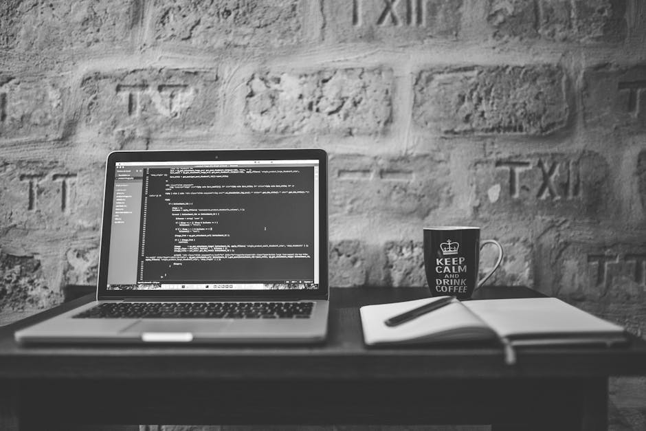 A black-and-white photograph of a workspace featuring an open laptop displaying lines of code on the screen, positioned on a dark wooden desk against a textured brick wall background. To the right of