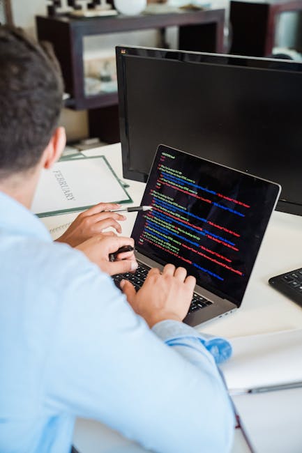A black-and-white photograph of a workspace featuring an open laptop displaying lines of code on the screen, positioned on a dark wooden desk against a textured brick wall background. To the right of