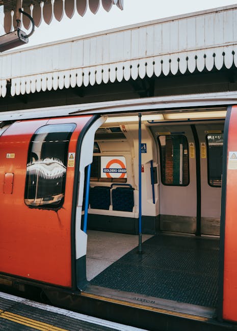 An open London Underground train carriage with a red exterior and a white interior, situated at a station platform under a white painted canopy with decorative scalloped edges. The platform surface is